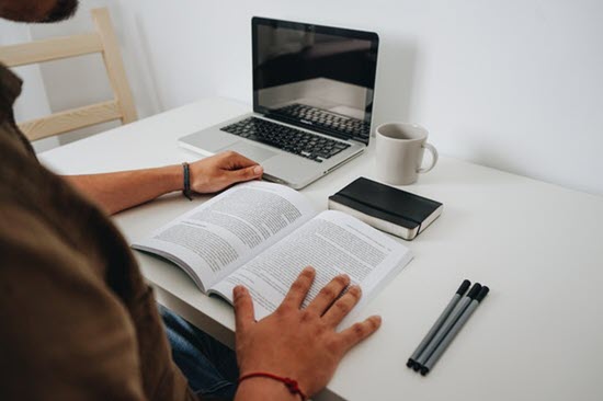 man sitting while reading a abook near his laptop